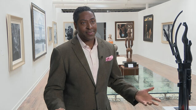 Gerald Griffin stands in a welcoming position with his arms open in the foreground of his gallery space which contains multiple paintings on the walls and sculptures on glass tables in the center.