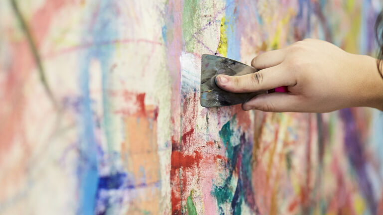 A zoomed in view of a person holding a palette brush against a large canvas with many swipes of colors such as red, turquoise, yellow, green, blue and orange. 