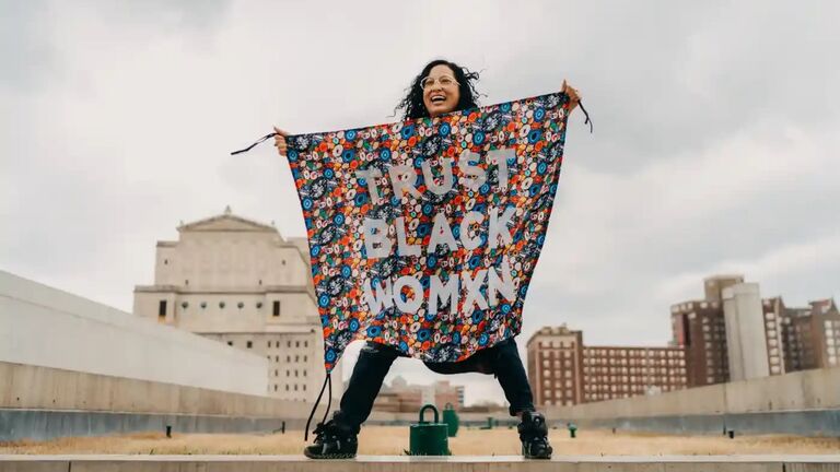 Artist Aram Han Sifuentes is holding up a colorful banner with the words “TRUST BLACK WOMXN” in white.