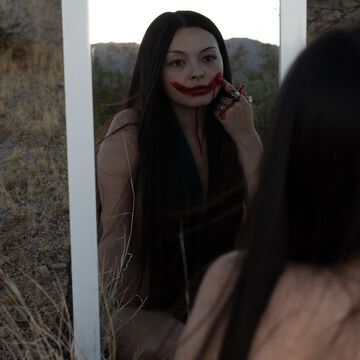 A person looks through a mirror as they draw a smile in blood-red paint across their face.