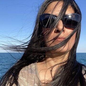 Gitanjali Kapila wears sunglasses and stands in front of a body of water, the wind blowing her hair across her face.