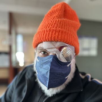 Joseph wears an orange knit hat, a navy blue medical mask and has one eye patched with tape and a what looks to be the metal lid of a glass parmesan shaker. 