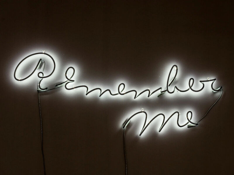 A neon sign spelling out Remember Me on a black wall