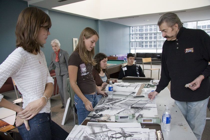 Students and faculty gather together around a table. On top of the table are many black and white drawings and sketches.