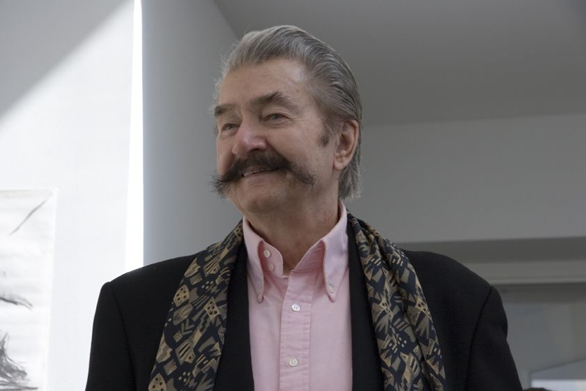 Leroy Neiman smiles while looking off to the side. He is wearing a pink button-up shirt, a black jacket, and a black and gold patterned scarf. He has grey hair and a full gray mustache.