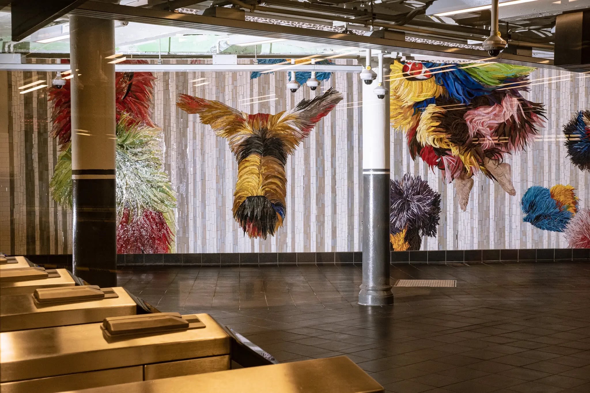 An image of Nick Cave’s colorful mosaic installation on the back wall of a subway station. In the left foreground, turnstiles are present.