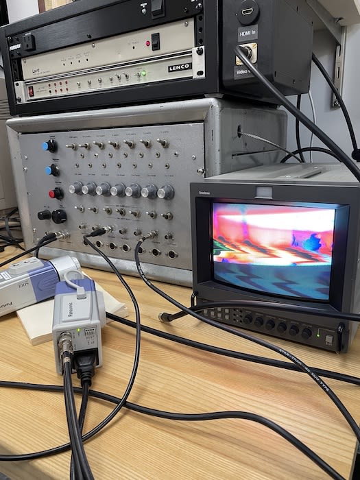 A large silver synthesizer on a wooden desk hooked up to a small television showing wavy lines of color