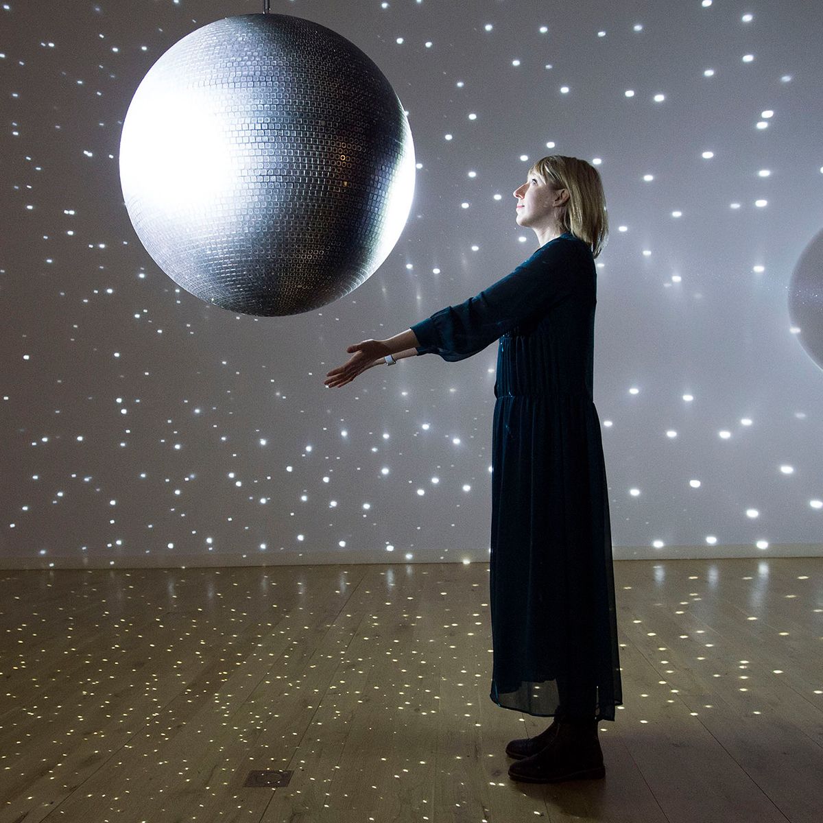 A photo of a female artist in a long dress with her arms outstretched in front of a large disco ball
