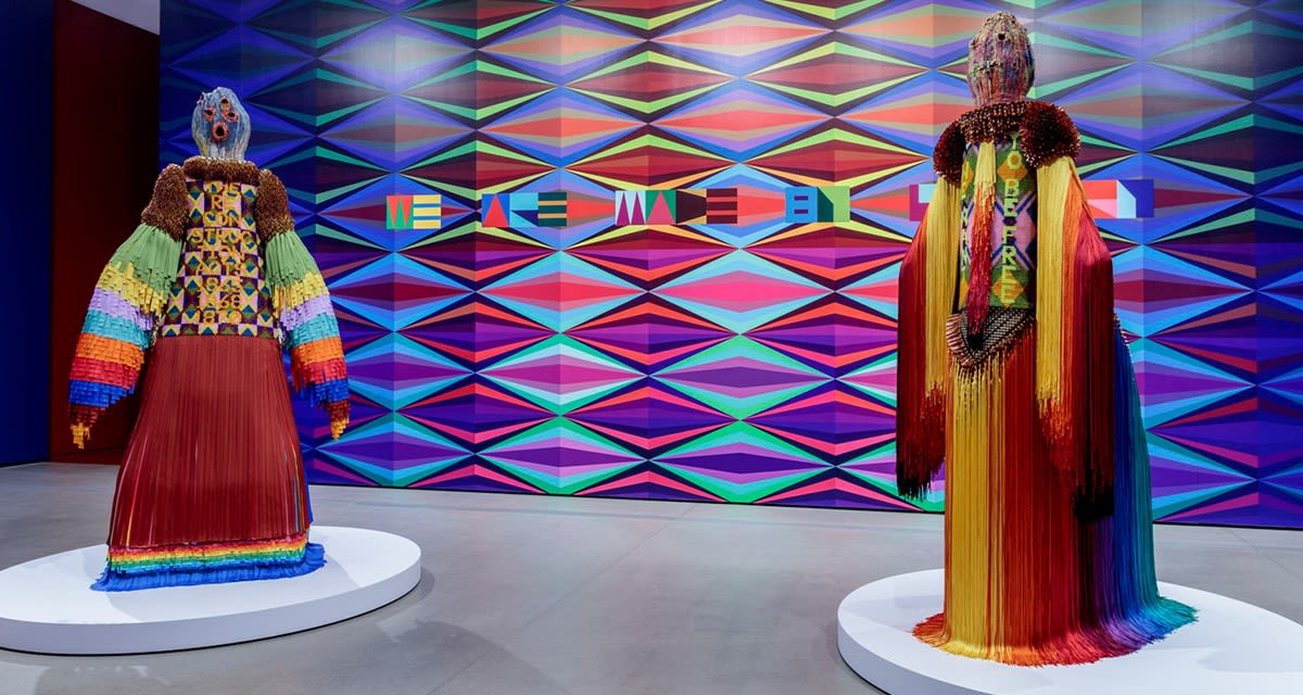 Two brightly clothed figures on display against a vivid geometric background