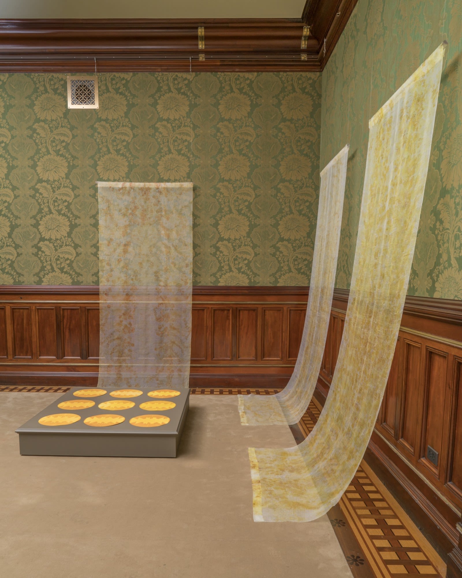 Textiles hanging in a room with green wallpaper and warm wood paneling