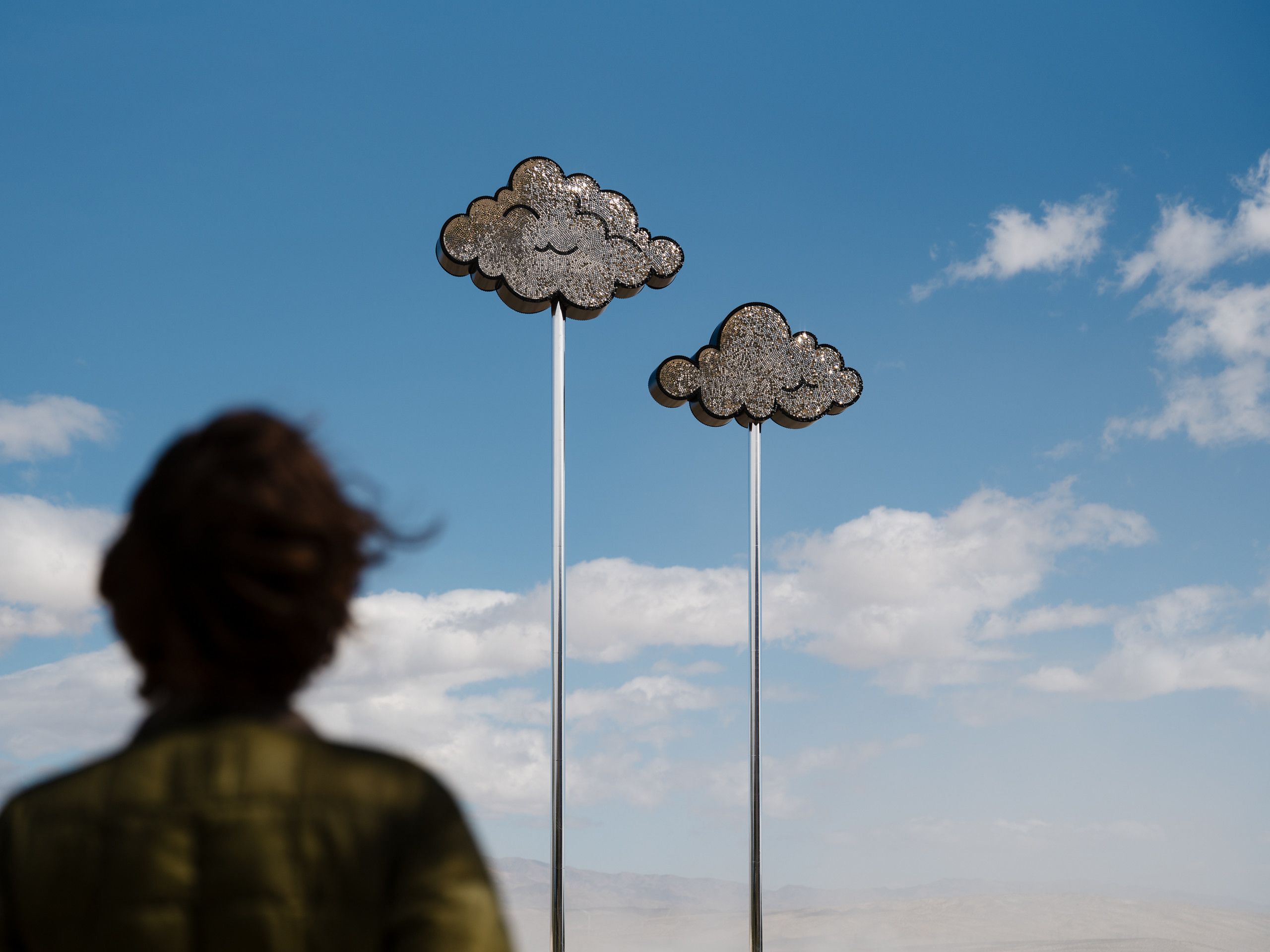 A figure stares towards two sculptures of silver cartoon clouds thrust into the blue sky
