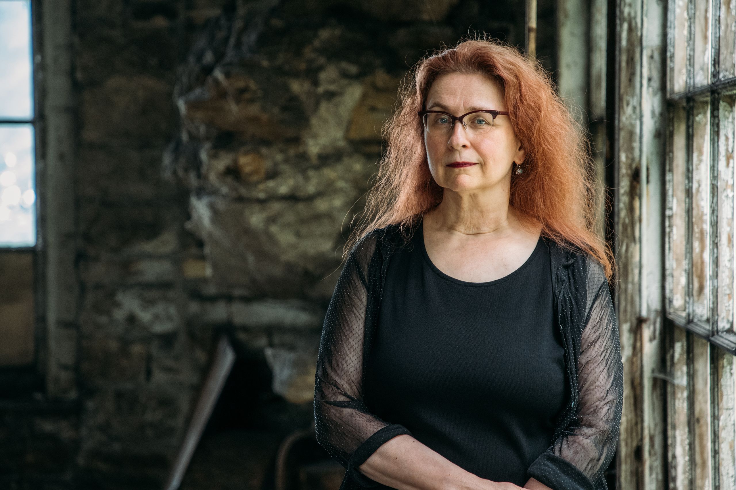 A red-haired woman in a black shirt stands inside a dilapidated stone-walled greenhouse