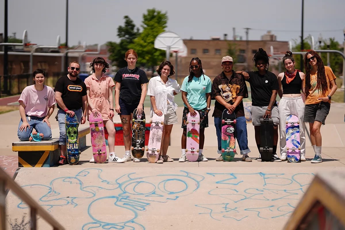 A photograph of of ten individuals looking at the camera and smiling. Eight of the ten people are holding skateboards. Several basketball hoops are in the background. Concrete flooring covered in blue spray paint is in the foreground.