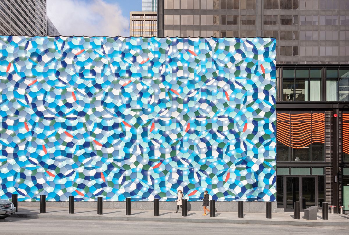 A huge, wavelike blue, green, and orange abstract painting stands on a Chicago street with skyscrapers in the background. A few people walk on the street in front, completely dwarfed by the painting