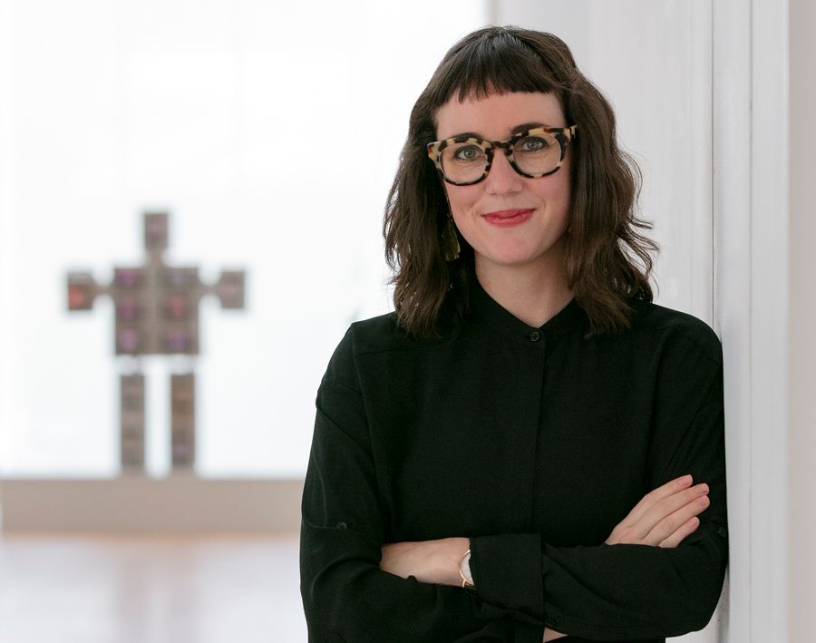 A white woman with dark brown hair and bangs cut straight across her forehead, wearing a black blouse and round, tortoiseshell glasses, crosses her arms, leans against a white all, and smirks at the camera. In the unfocused background is a robot-like sculpture.
