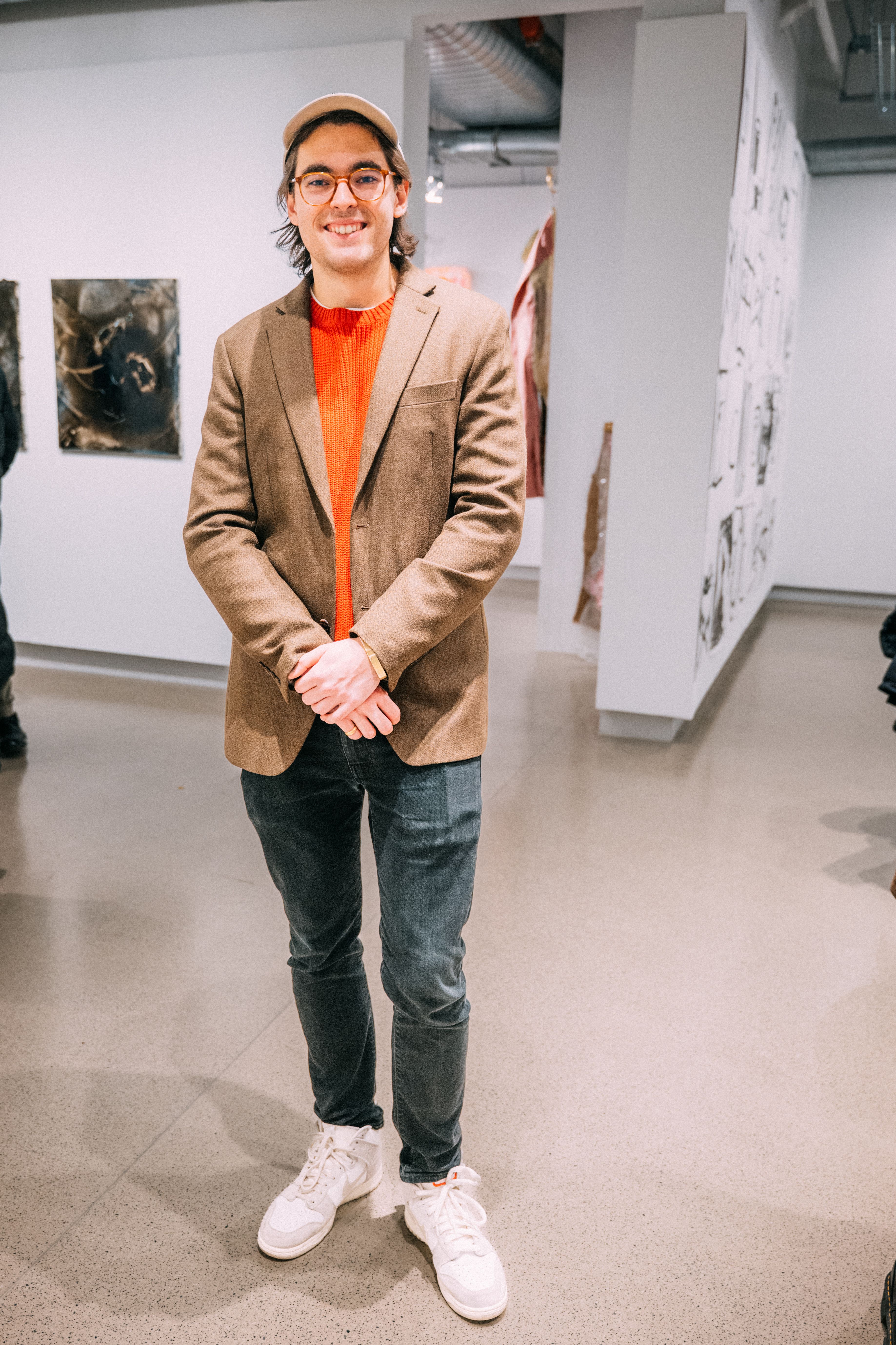 A student poses in the gallery while wearing an orange sweater, brown jacket, jeans, and white Nikes.