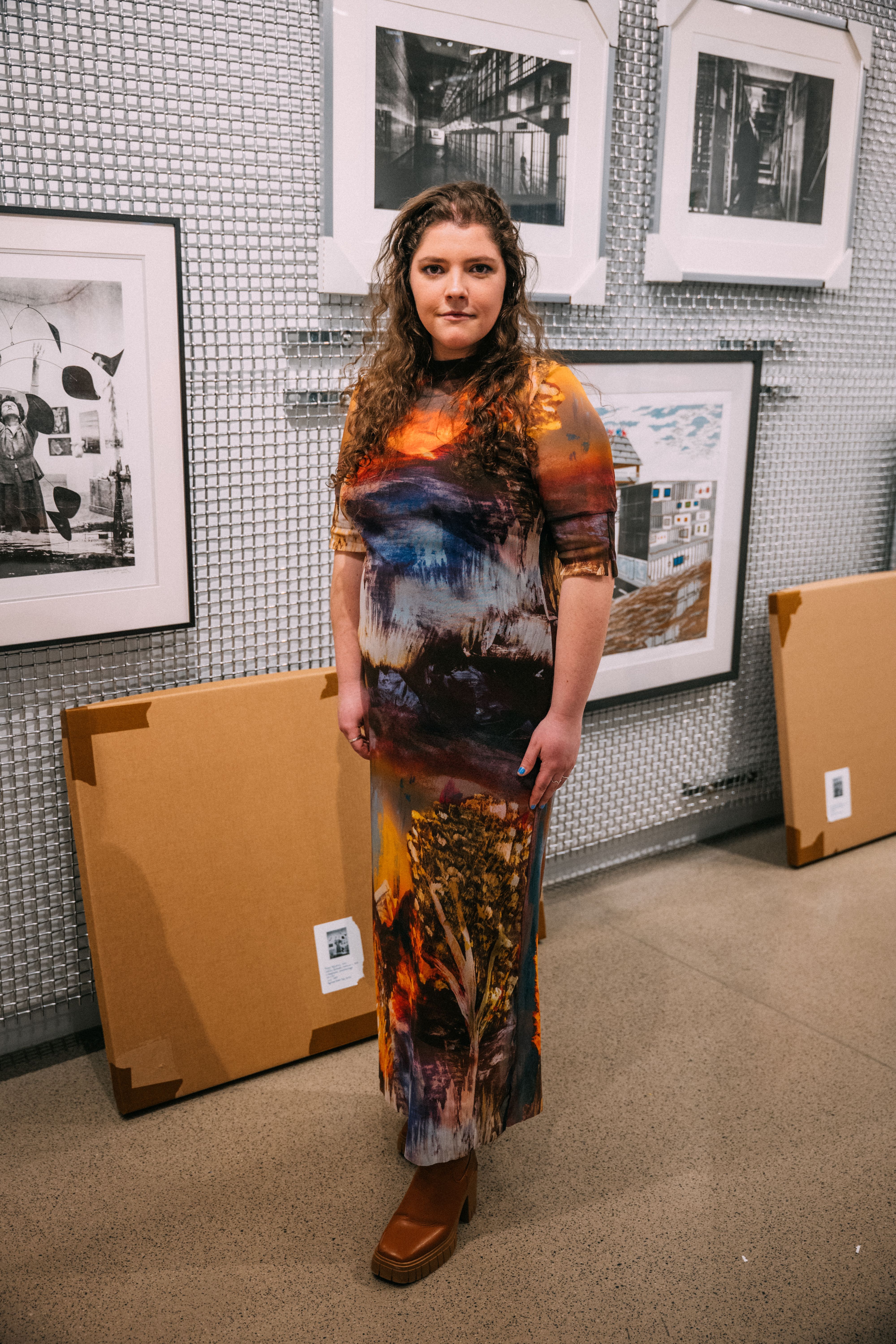A student stands in front of a wall of framed artwork while wearing a long dress in orange, blue, and brown shades.
