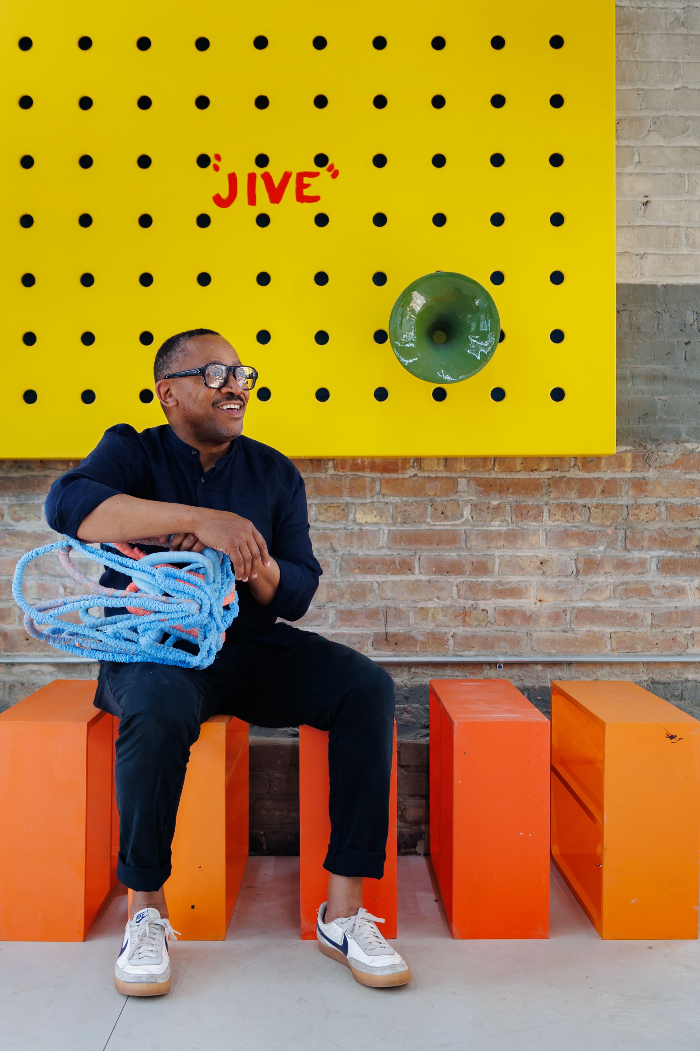 Norman Teague sits on an orange bench in front of a yellow peg board painted with the word "JIVE"
