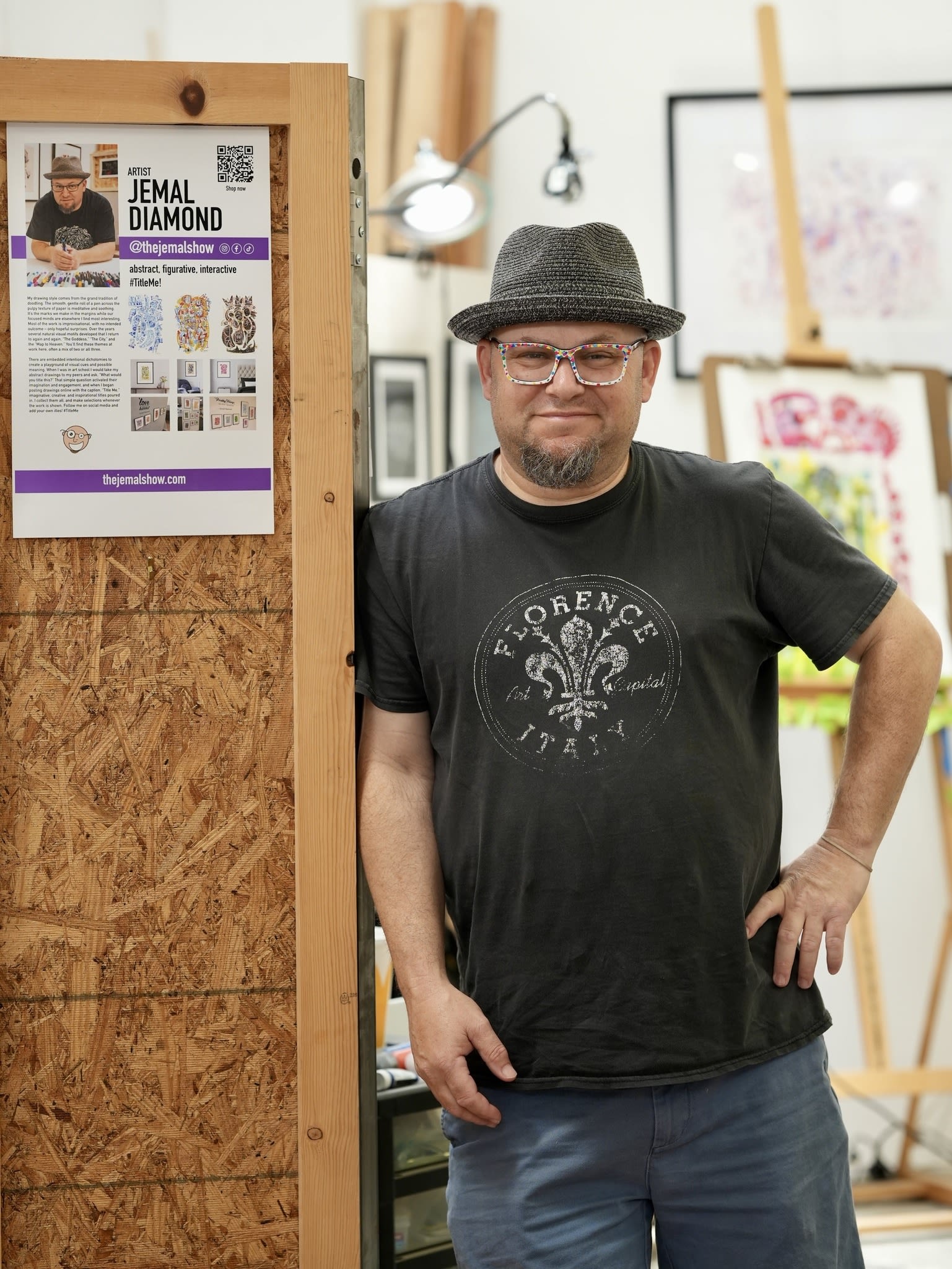 A person in a hat and glasses poses in their studio