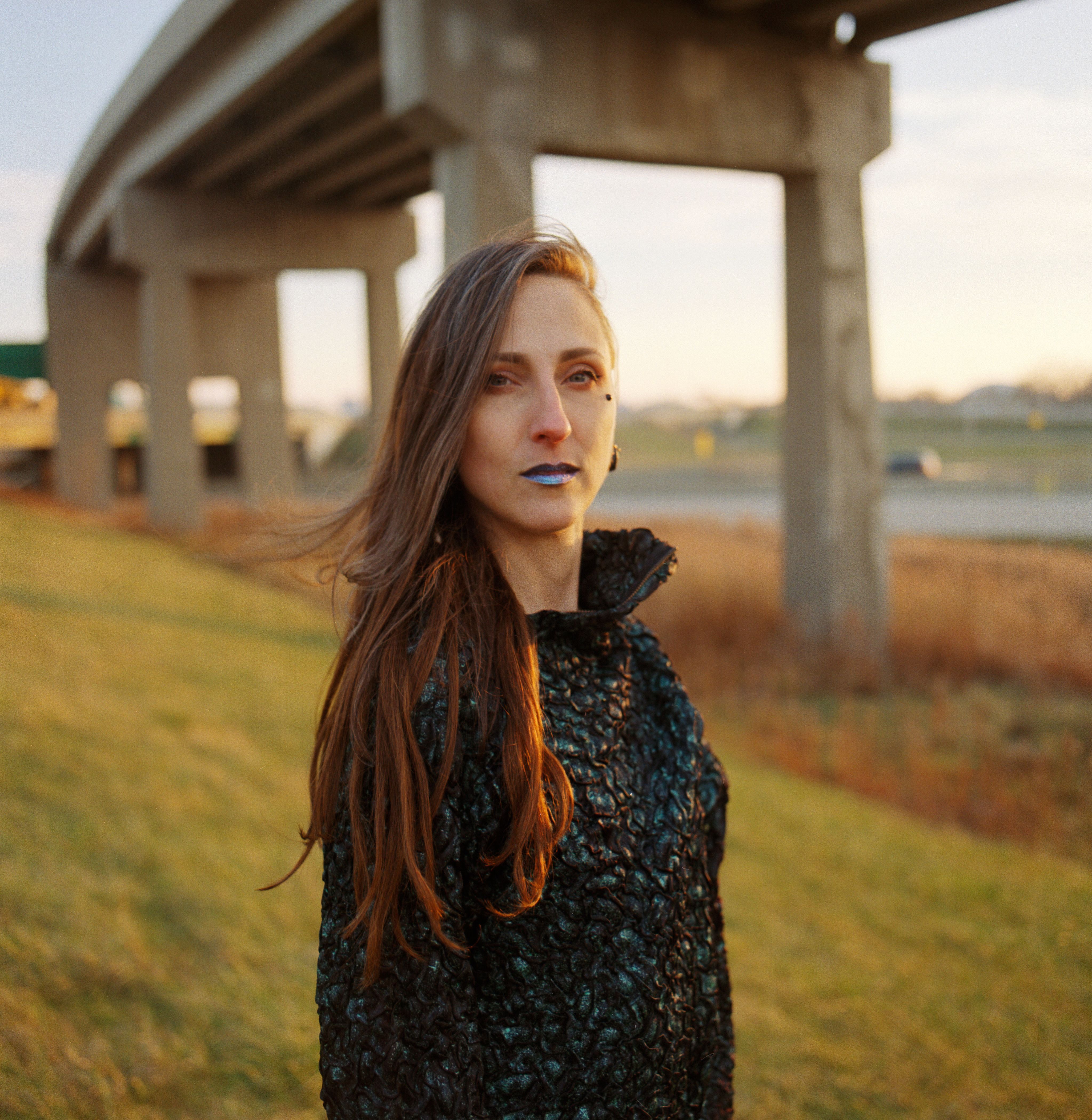 An artist in a sculptural black top and metallic blue lipstick stands in a field under a freeway