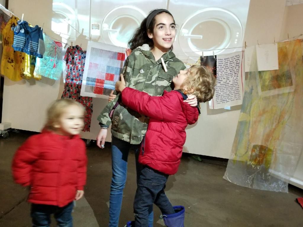 Three children stand in front of a clothespin strung with artwork. 