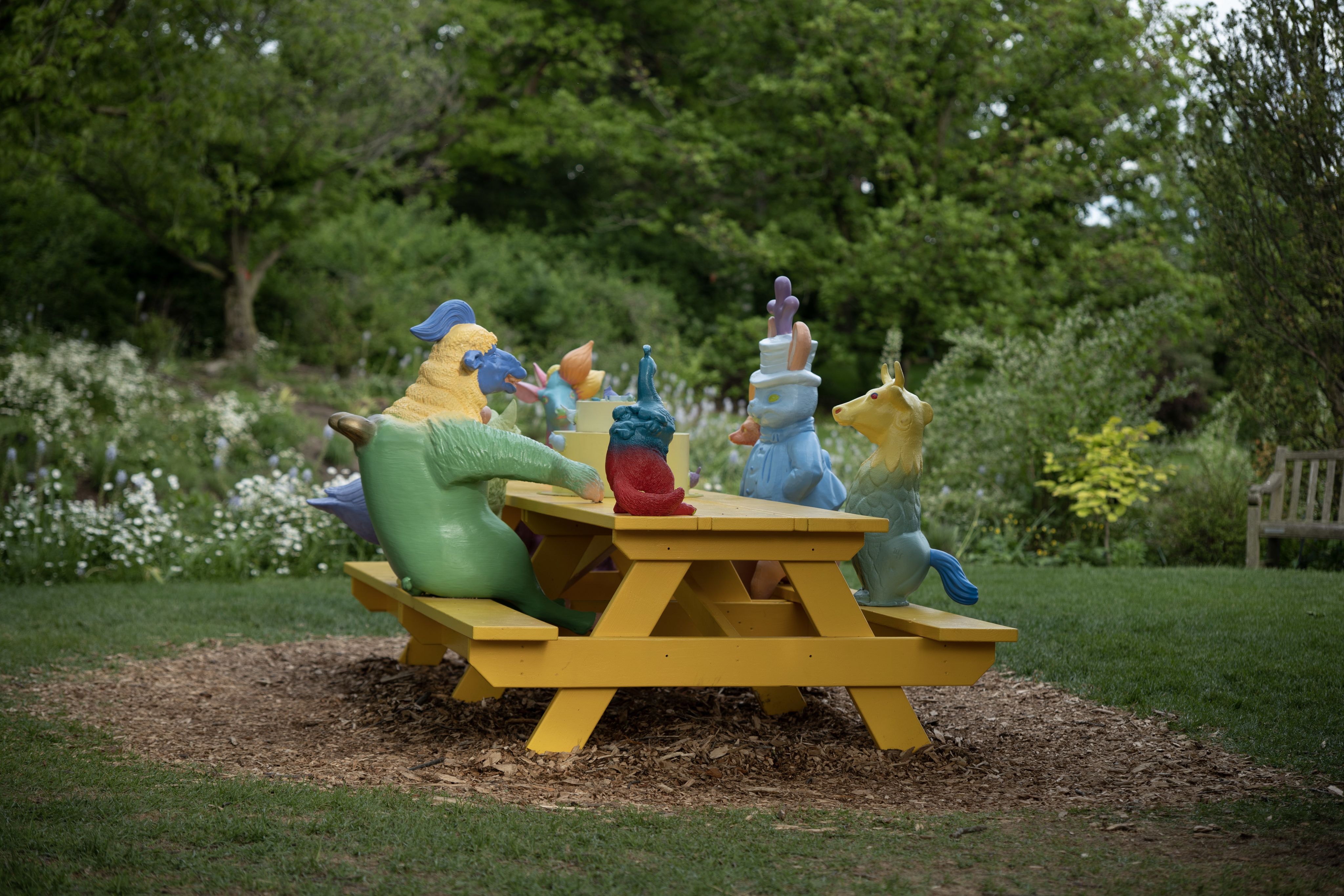 A photograph of colorful animal sculptures sitting at a yellow picnic table. A yellow cake sculpture sits in the middle of the picnic table.