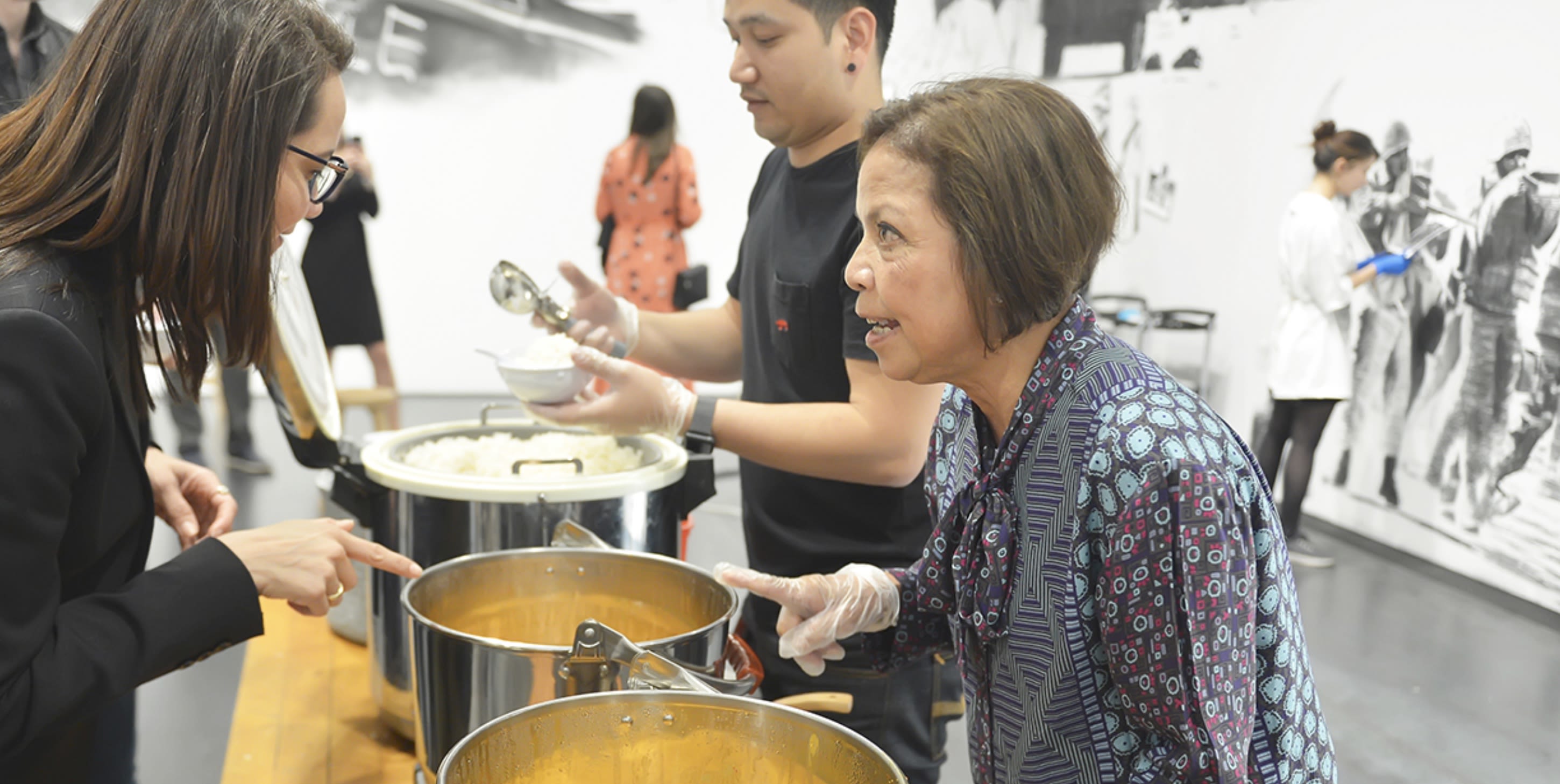 An image of Rirkrit Tiravanija's performance (who's afraid of red, yellow, and green). The image features people serving others rice and curry. In the background, people view the artwork on the walls.