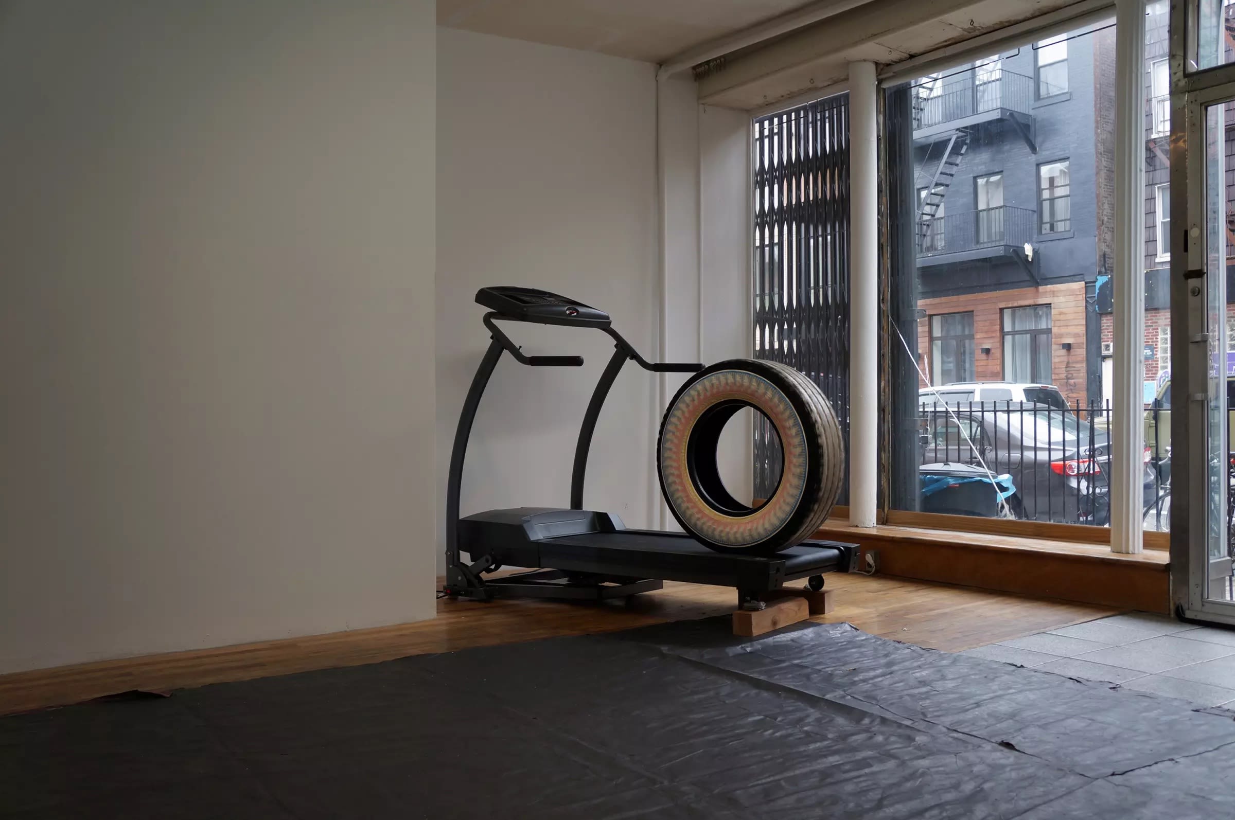 A photograph of an installation by Gregory Bae. The installation piece is black and shaped like a treadmill with a large tire at the end of it.