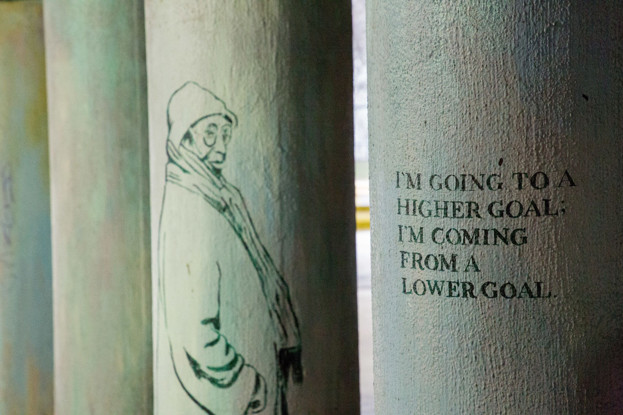 A mural painted on columns under a train overpass, the text of which says "I'm going to a higher goal; I'm coming from a lower goal"