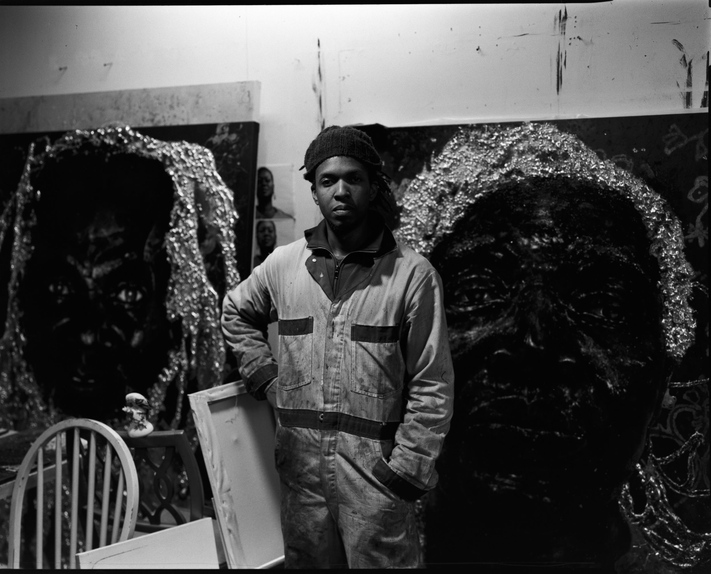 A black and white photo of a man in a jumpsuit standing in his studio in front of paintings