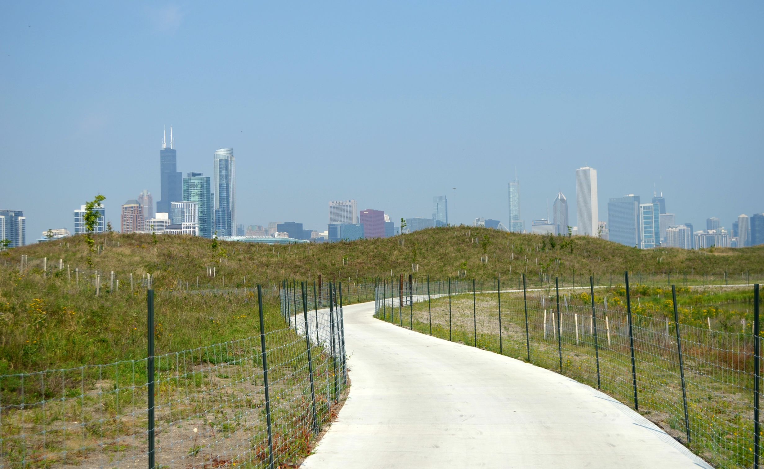 A concrete path winds through a green space, with the city of Chicago skyline in the background