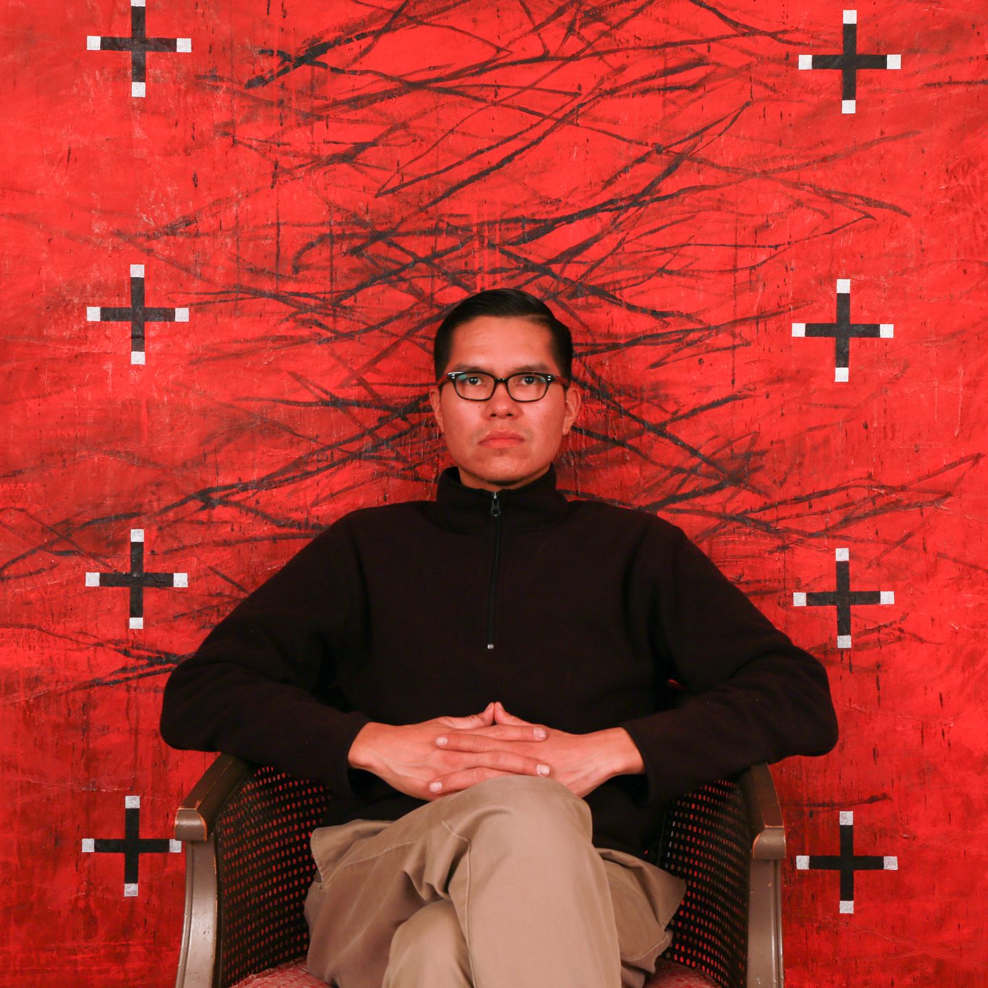 A man in glasses sits on a chair posing in front of a large, saturated red and black painting