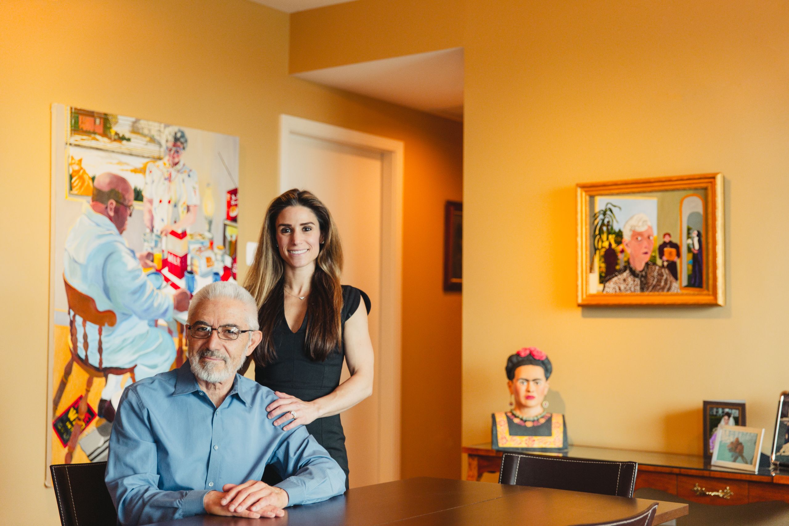 An older man and his daughter pose in a yellow-colored room in their house. A painting of a man sitting at a table is behind them