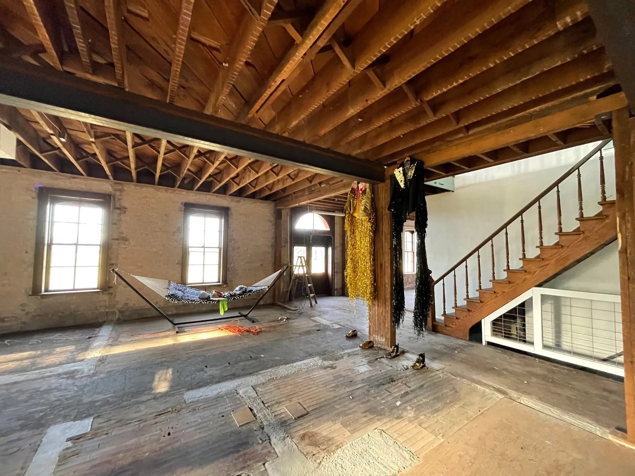 An interior view of the Poor Farm. The space is bare and wooden, with only a hammock, a ladder, and two hanging costumes in the room.