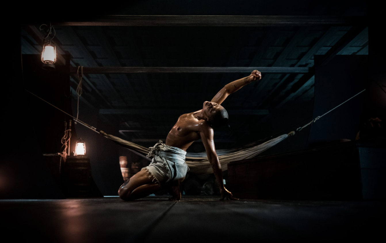 A shirtless man kneels down on the lower level of a ship with his right arm stretched over his head.