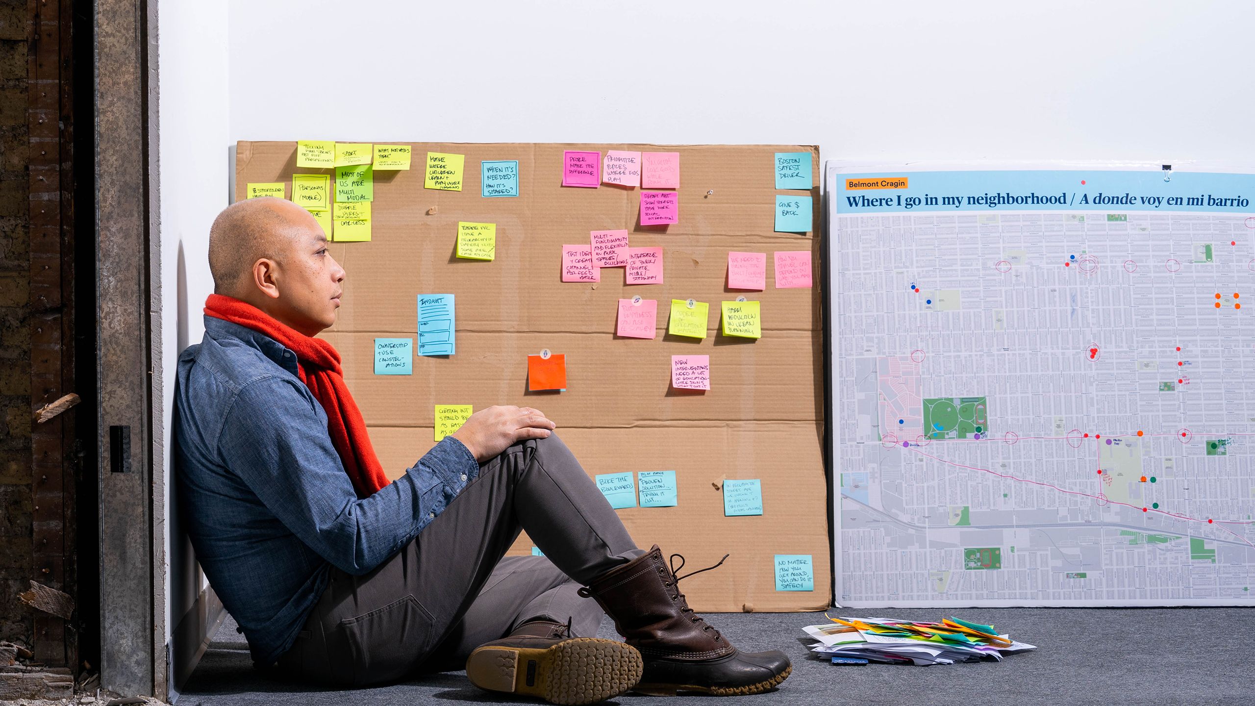 A man in a red scarf and blue shirt sits on the floor of an office space next to a cardboard board with colorful post-its and a neighborhood map