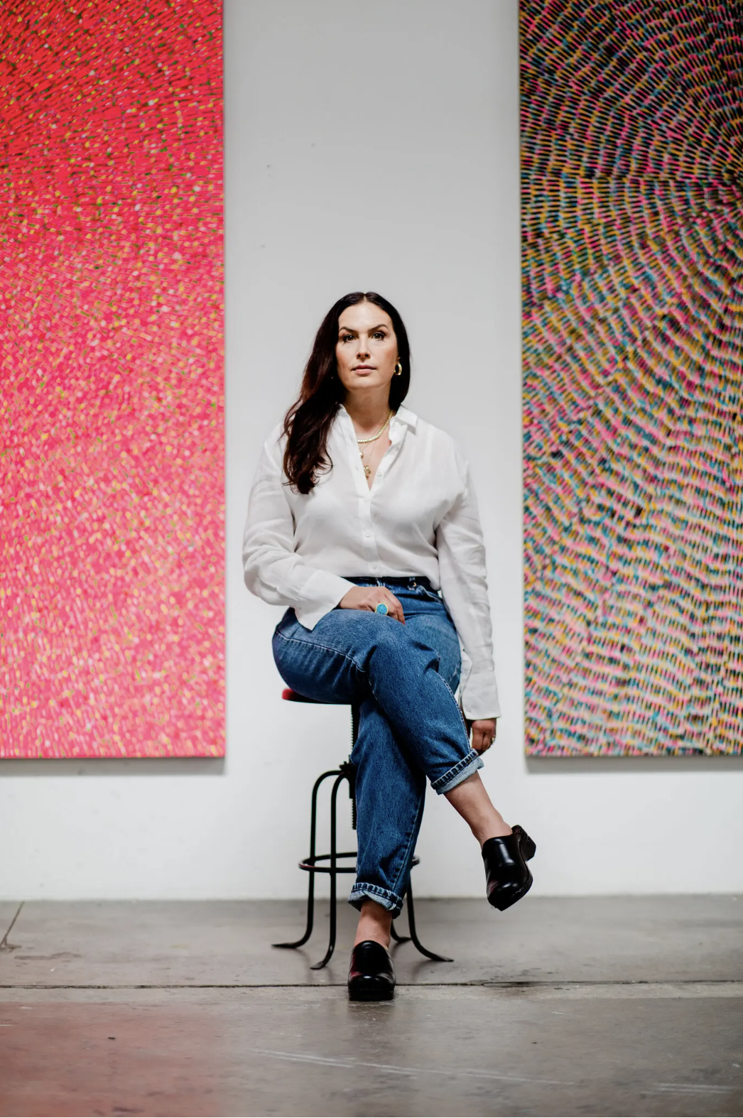 A woman with long, brown hair sits on a stool in front of two large piece of artwork that hang side-by-side on a white wall. The woman is wearing a long-sleeve white button-up shirt and blue jeans. The artwork on the left is light-red but is made of many dots that are different colors. The artwork on the left shows a multicolored circular pattern that radiates from the center of the canvas out towards the canvas' edges.