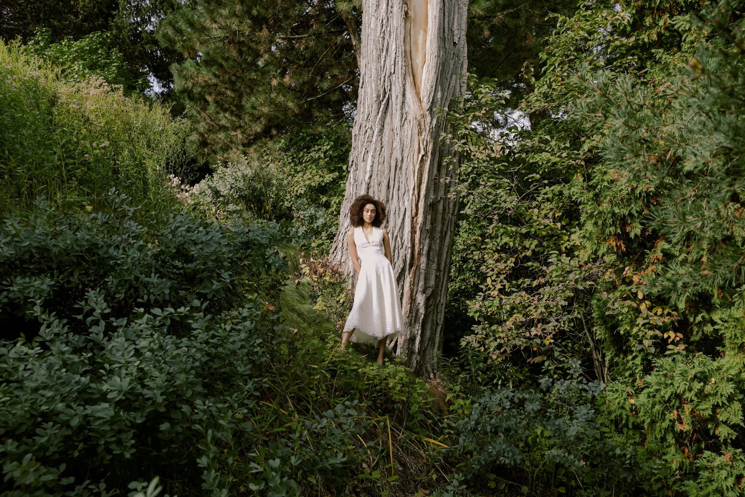A young woman in a white dress stands in front of a tree in the woods
