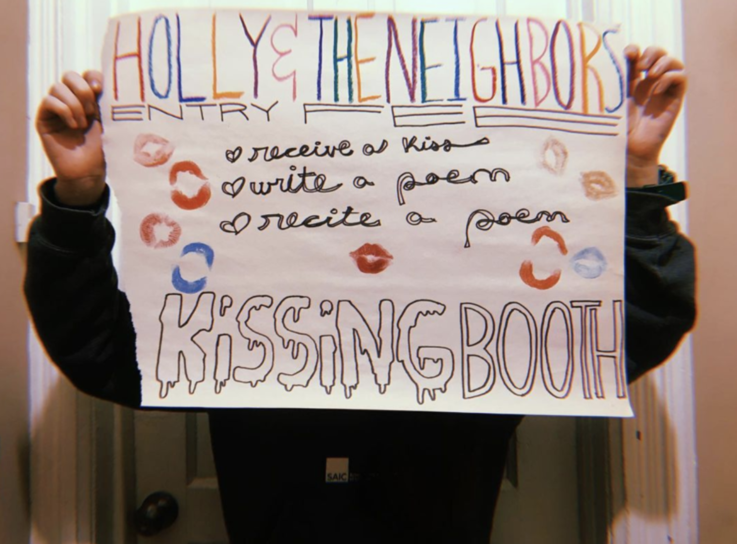 Someone holds up a colorful poster that obscures their face. The poster is decorated hearts, multi-colored lipstick stains, and multi-colored letters advertises an event called "Kissing Booth," and event where people can "receive a kiss, write a poem," and "recite a poem," as is written on the poster.