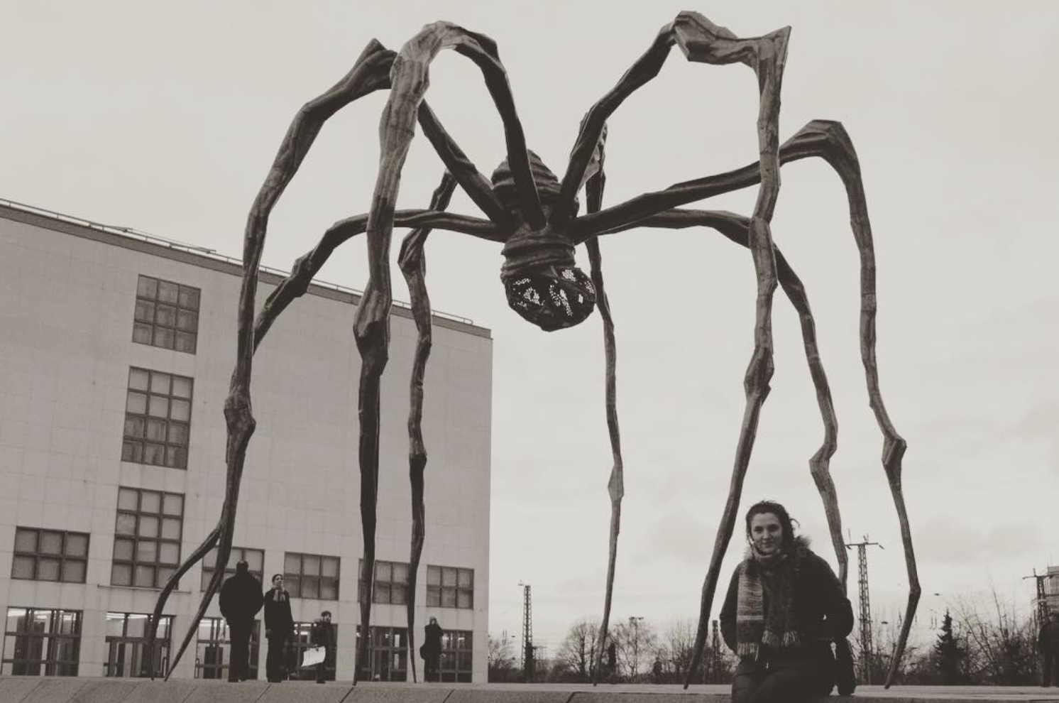 A large sculpture resembling a spider stands in front of a building. A person poses in front of the sculpture for a picture.