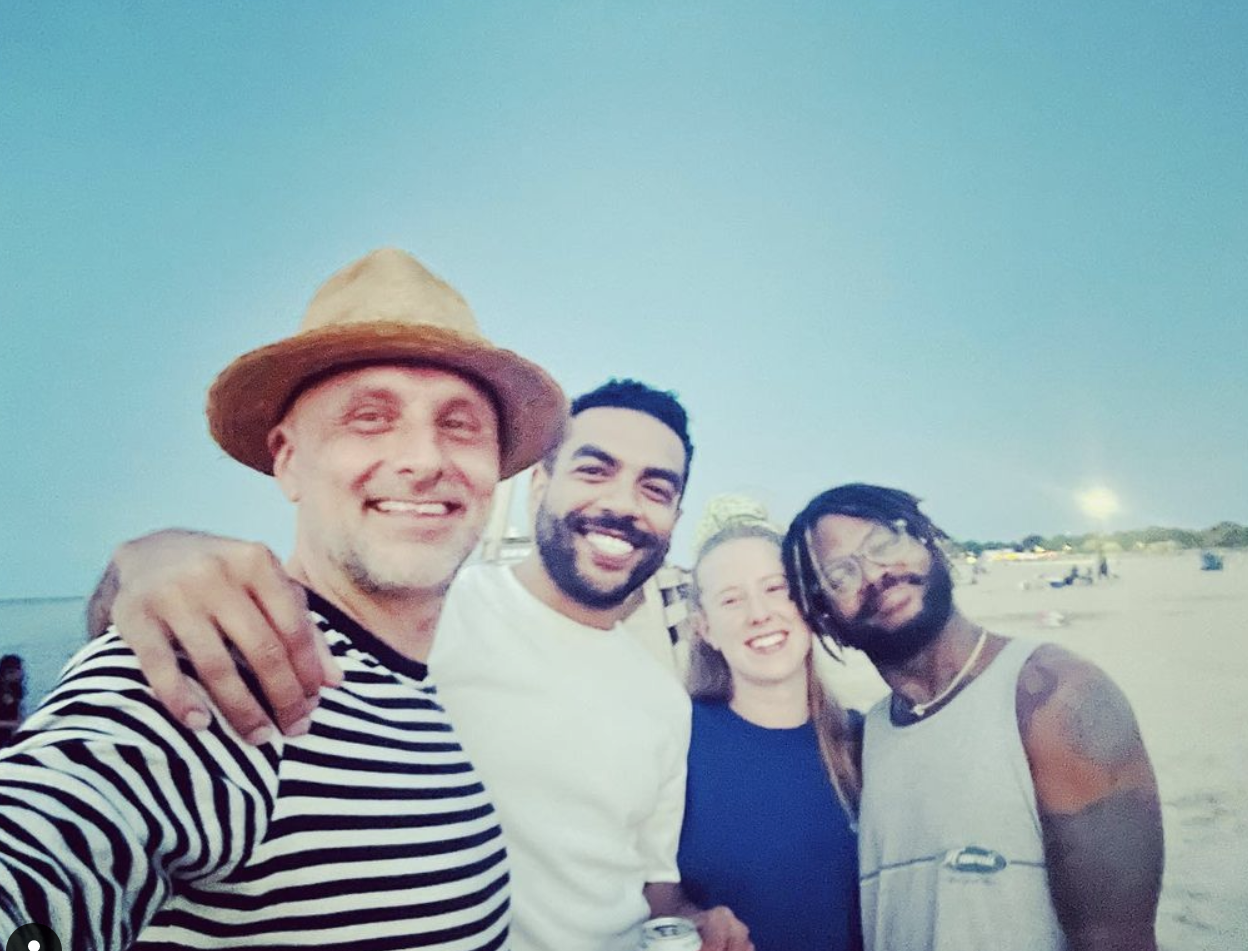 A group of four people pose for a selfie while on the beach. They smile as they wrap their arms around each other's shoulders.