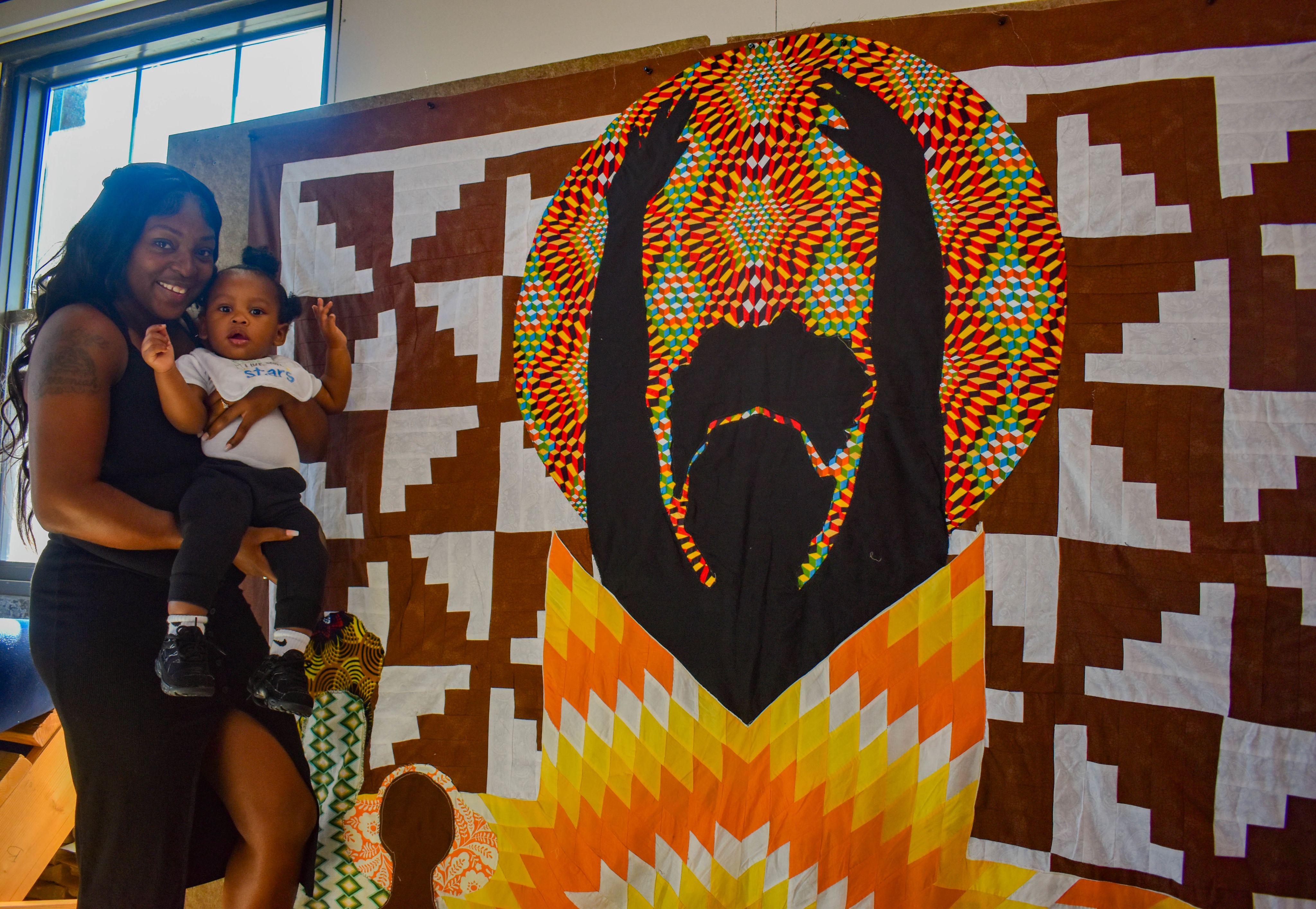 A mother and child stand before a quilt