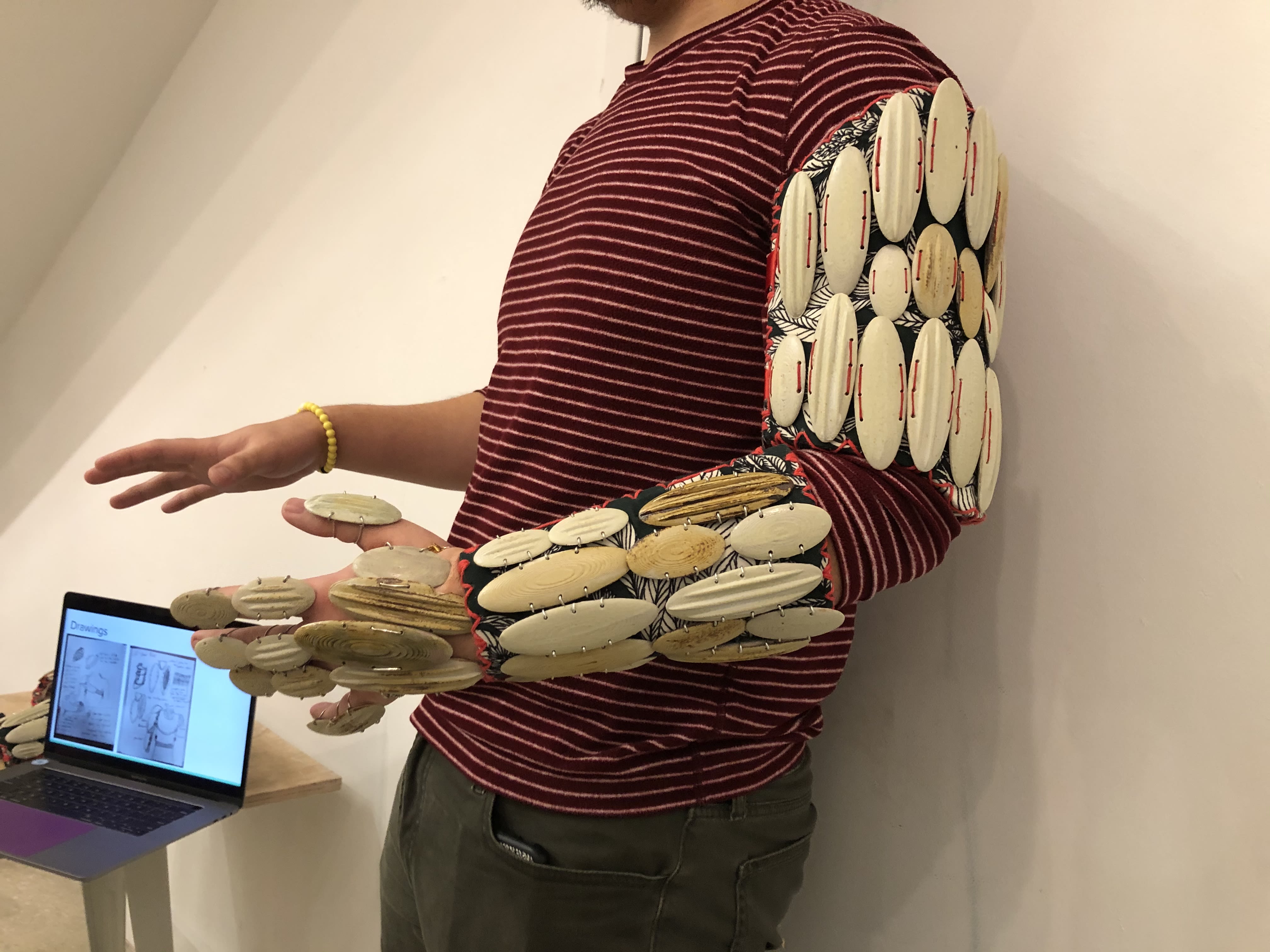 A student wears armor made of high-fired porcelain oval plates over the length of their left arm created in the style of Chinese jade armor. An open laptop sits on a table behind the student.