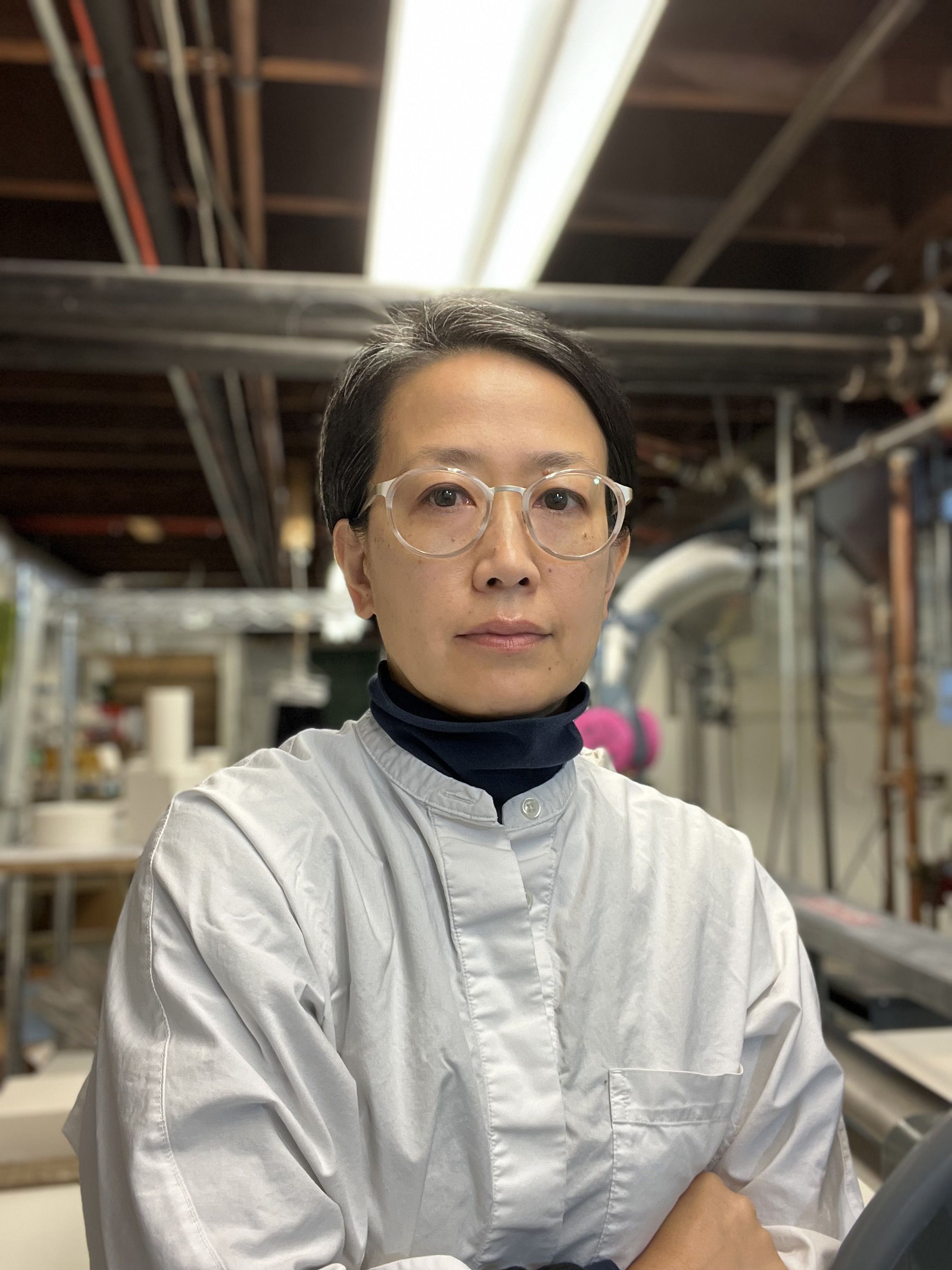 Mie Kongo faces the camera while crossing her arms. She is wearing a gray shirt and glasses with clear frames. Her studio can be seen in the background.