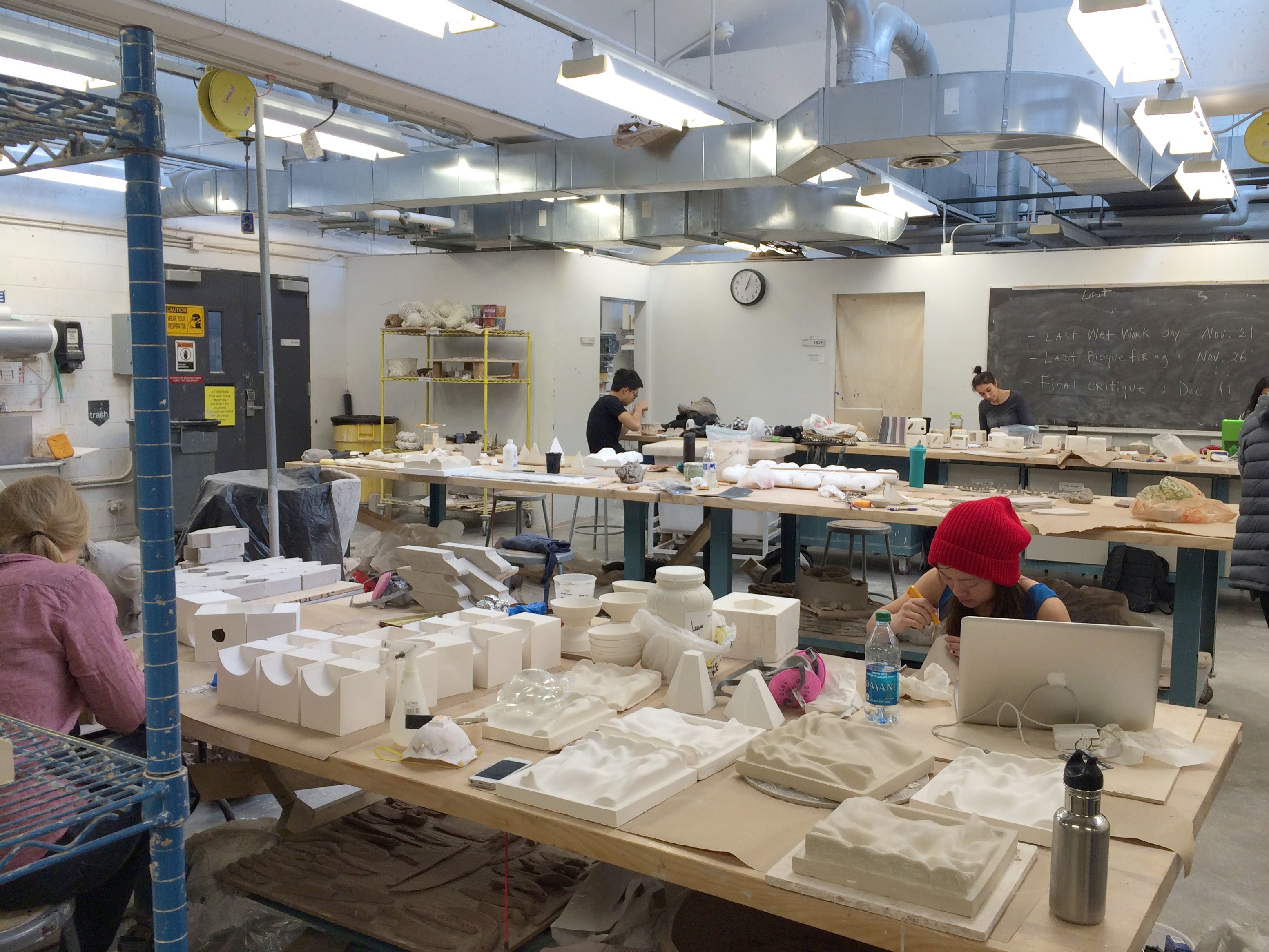 A classroom contains many large wooden tables. On the tables are plaster molds of various shapes and sizes. A few students are sitting down and working on the molds in front of them.