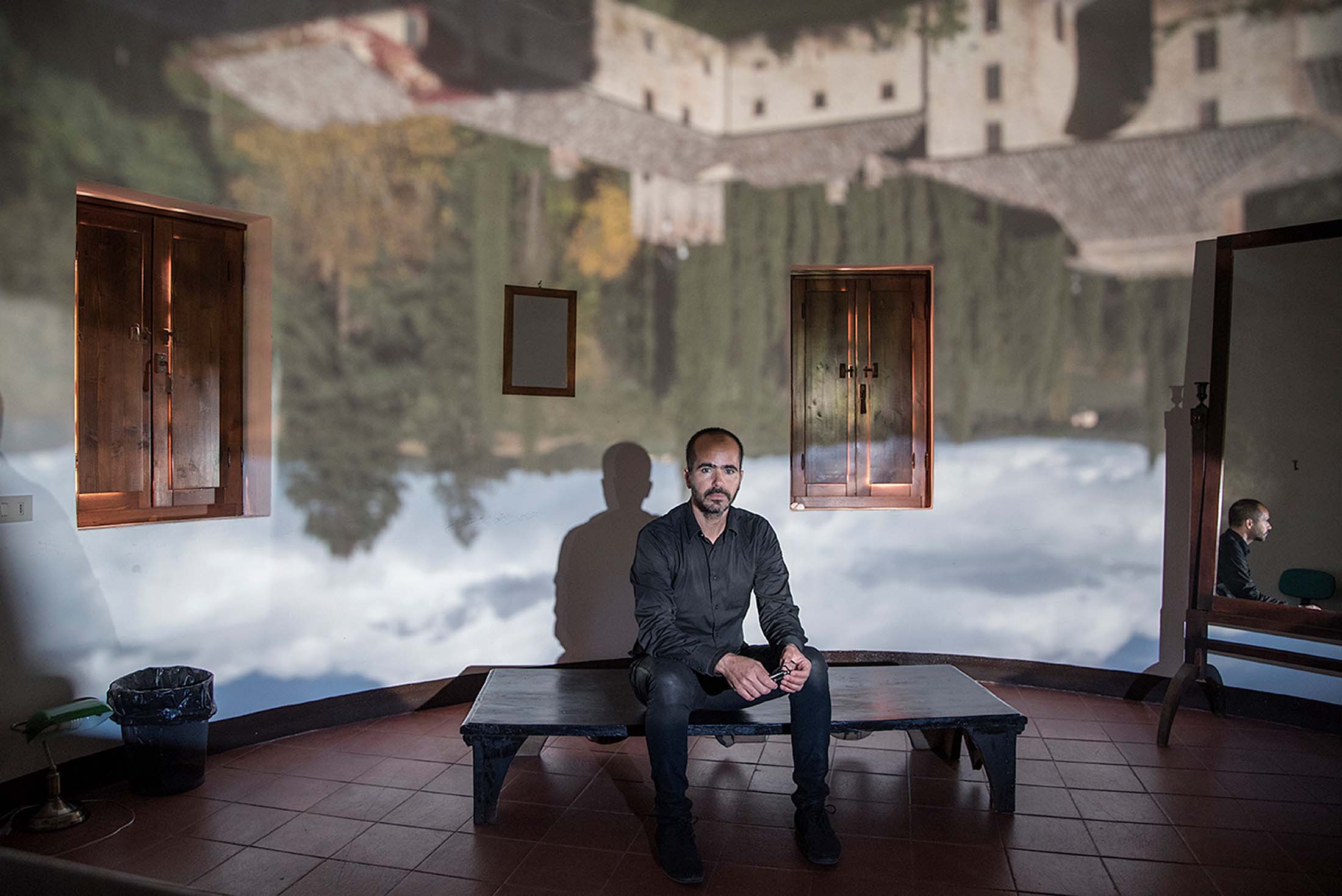 A man sits on a bench in a bare room with a sky and forest projected onto the walls