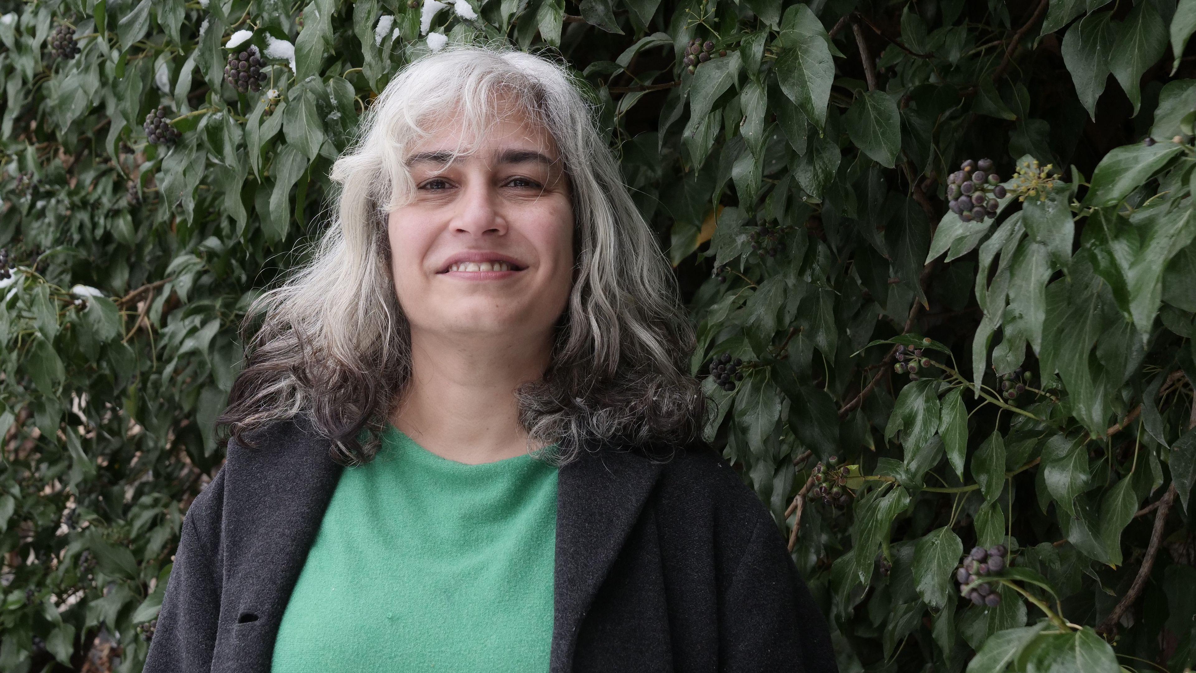 A woman with white and black hair stands in a green shirt and black blazer in front of greenery