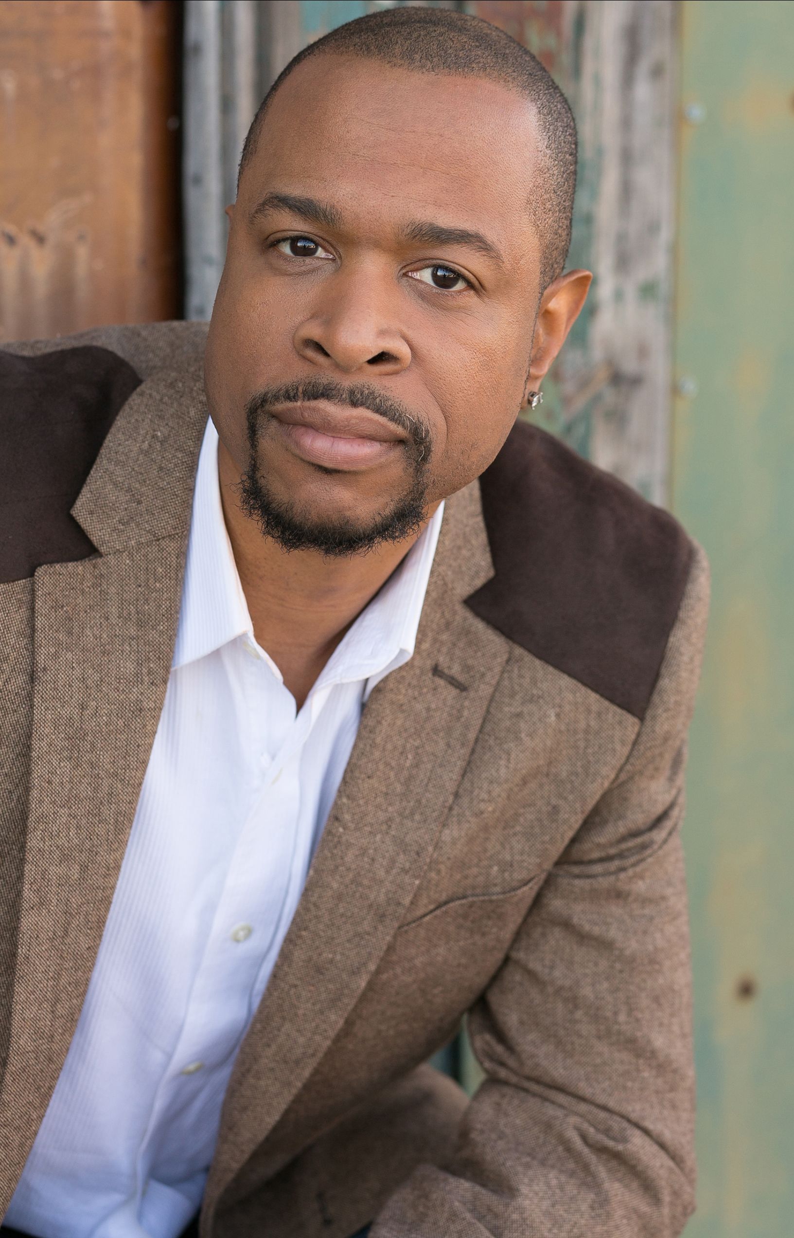 A Black man with a goatee wearing a white button-up shirt and two-toned brown blazer looks into the camera and smirks