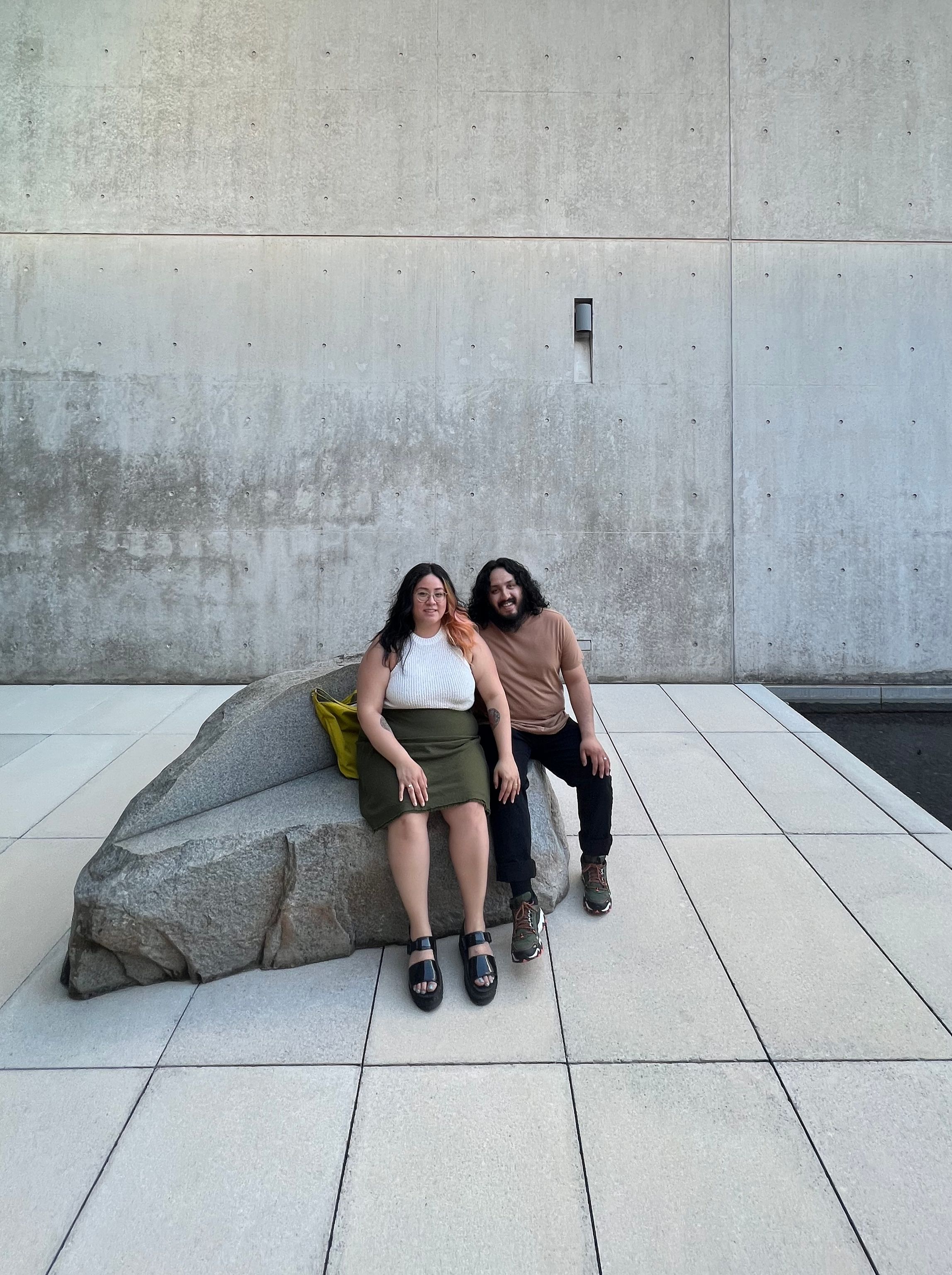 A couple sit on a stone bench in front of a concrete wall