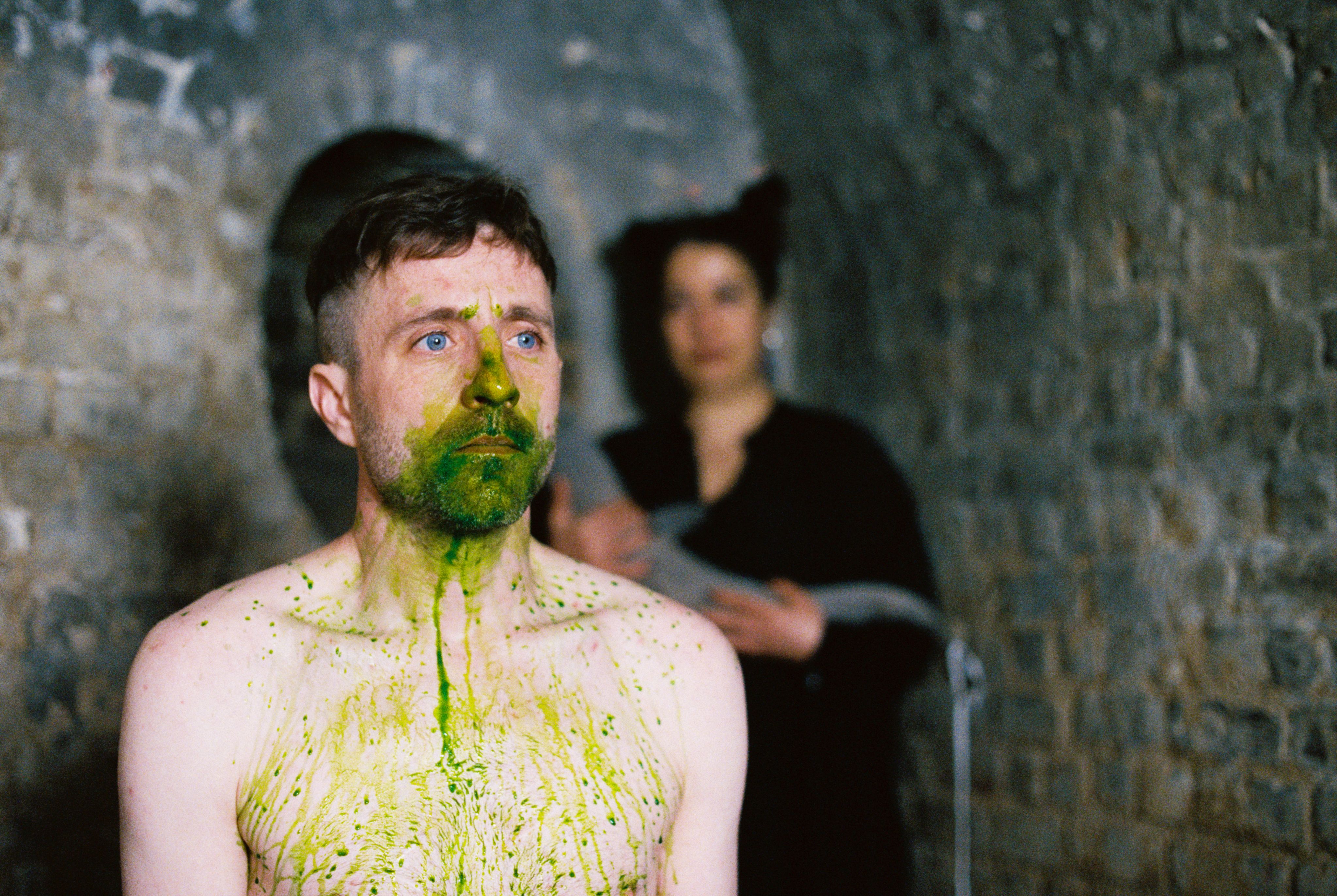 An artist with green paint on his face stands in a brick tunnel